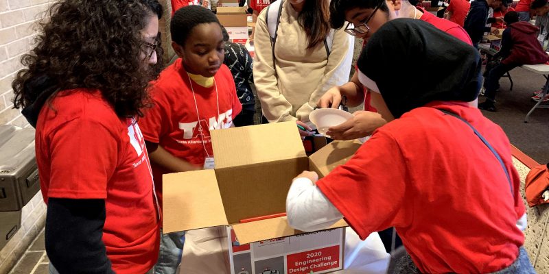 A diverse team of five students in red TAME shirts look into a box while building a prototype in the 2020 Gulf Coast STEM Competition.