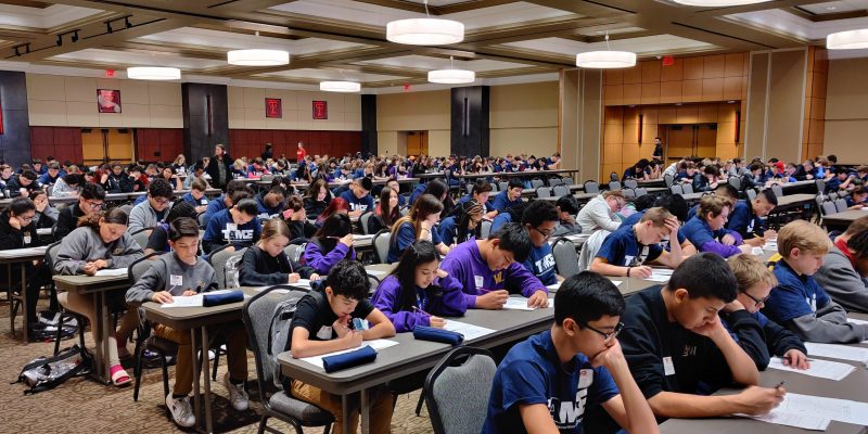 A diverse group of students wearing TAME shirts, grades 6-12, are pictured taking tests in a large auditorium with the Texas Tech University Logo on the walls.