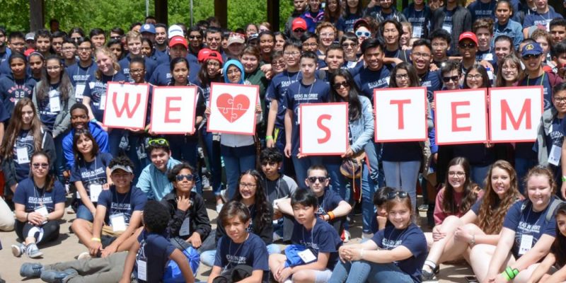 Large group of TAME students pose for a photo in a green field. Students in the middle hold signs that spell out We Heart STEM. From the 2018 State STEM Competition.
