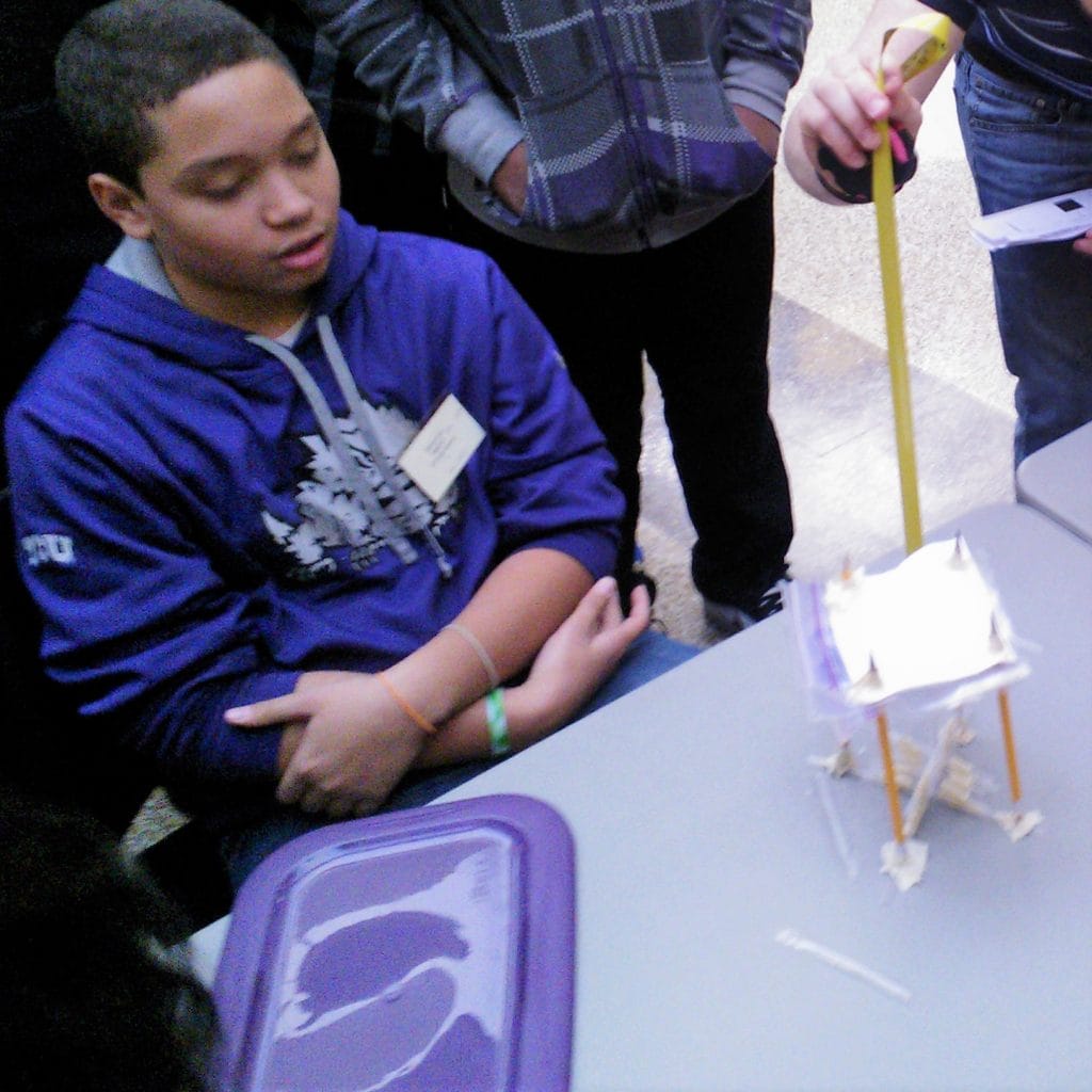 A student looks on as a volunteer Engineering Design Challenge Judge tests a student prototype at the 2014 Dallas Divisional Math & Science Competition, hosted at Tarrant County College.